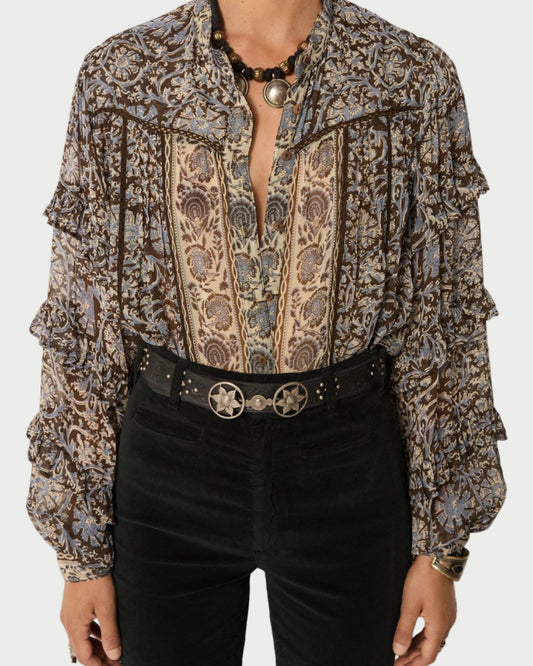 A person wearing the Maison Hotel Camilla Blouse—patterned, long-sleeved, with blue and brown florals—paired with black high-waisted pants, a black studded star-buckle belt, and a beaded necklace.