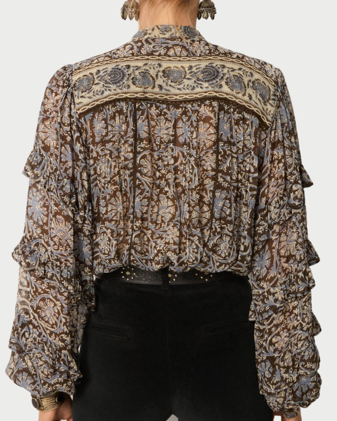 A person wearing the Maison Hotel Camilla Blouse—a sheer, long-sleeve viscose georgette top with a brown, beige, and blue floral pattern and ruffled sleeves—paired with high-waisted black pants. Photo taken from behind.