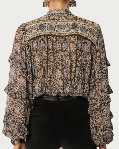 A person wearing the Maison Hotel Camilla Blouse—a sheer, long-sleeve viscose georgette top with a brown, beige, and blue floral pattern and ruffled sleeves—paired with high-waisted black pants. Photo taken from behind.