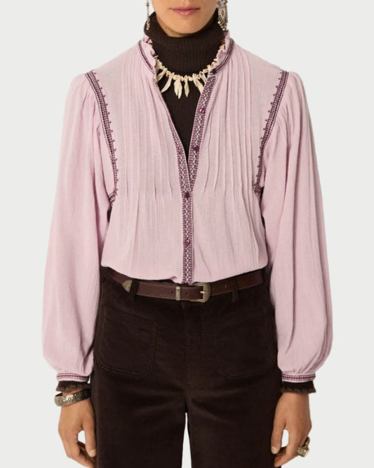 A person wearing the Maison Hotel Lila Blouse in light lavender cotton crepe with pleats and embroidery by Maison Hotel, layered over a dark turtleneck, paired with black pants, a belt, bracelets, and a shell necklace.