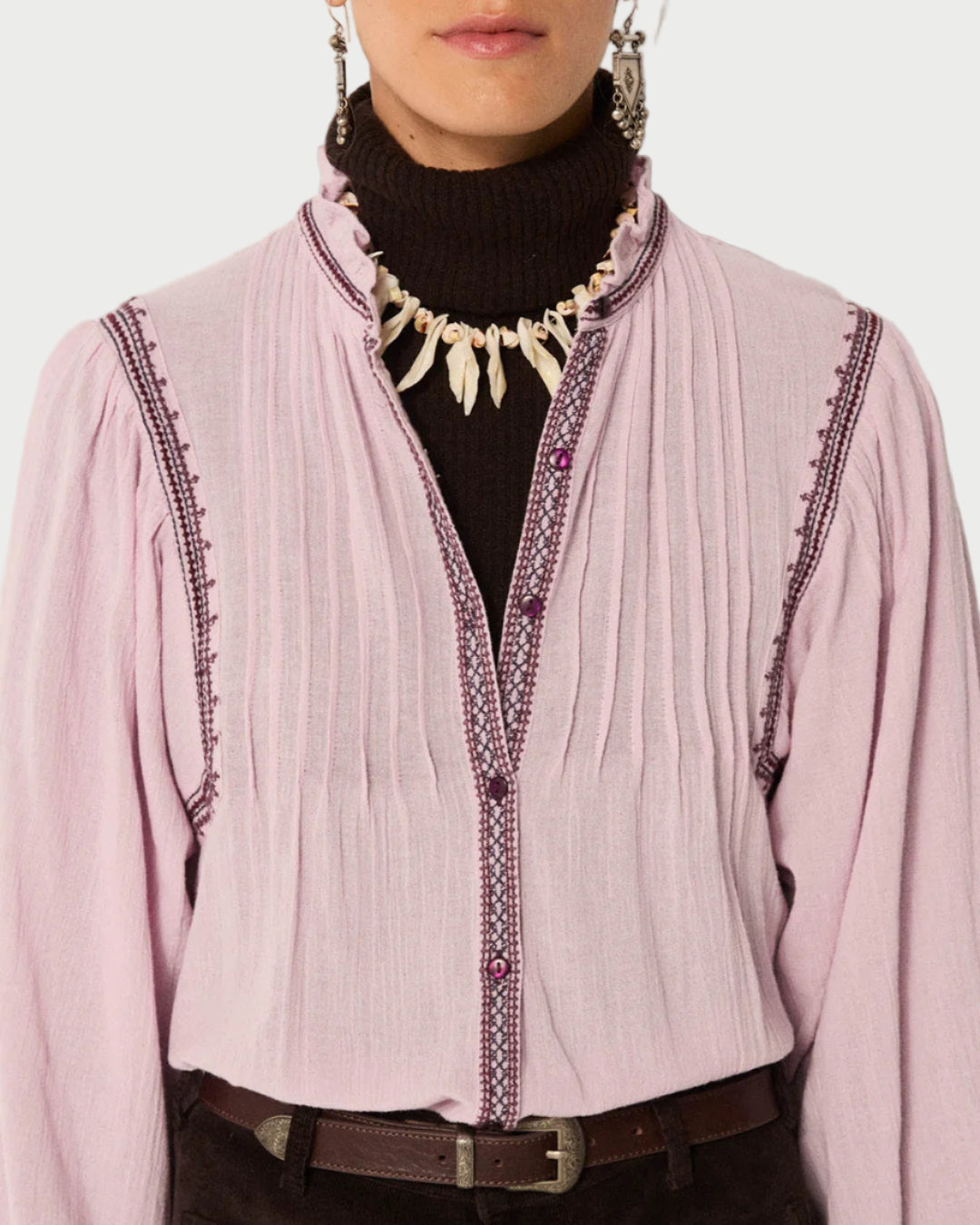 A woman wears the Maison Hotel Lila Blouse by Maison Hotel—a light pink cotton crepe top with decorative stitching—over a black turtleneck, paired with a white pendant necklace and dangling earrings. The photo is cropped from shoulders to waist.