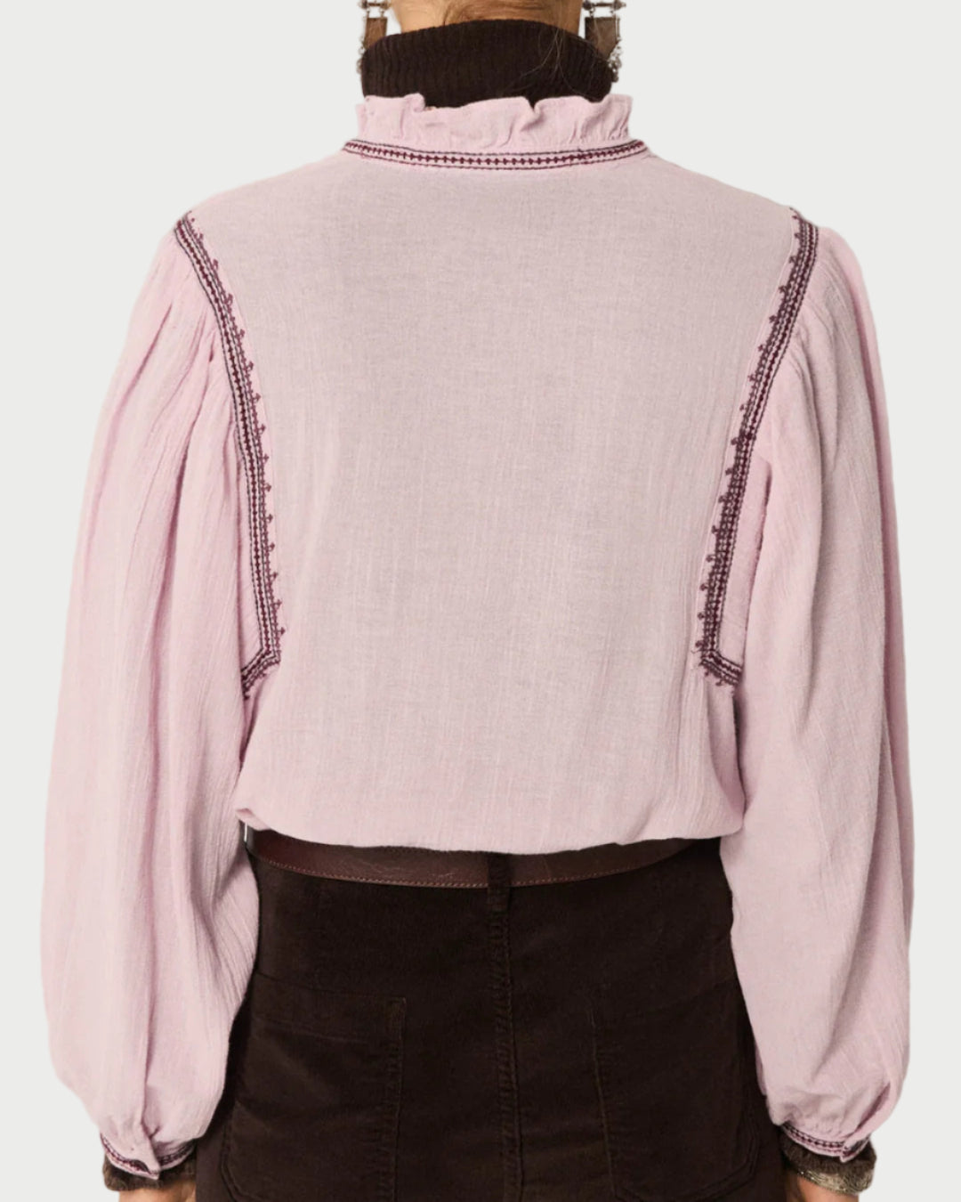 A person is seen from the back wearing the Maison Hotel Lila Blouse by Maison Hotel—a light pink cotton crepe top with gathered sleeves and dark decorative stitching at the shoulders—tucked into a dark skirt or pants.