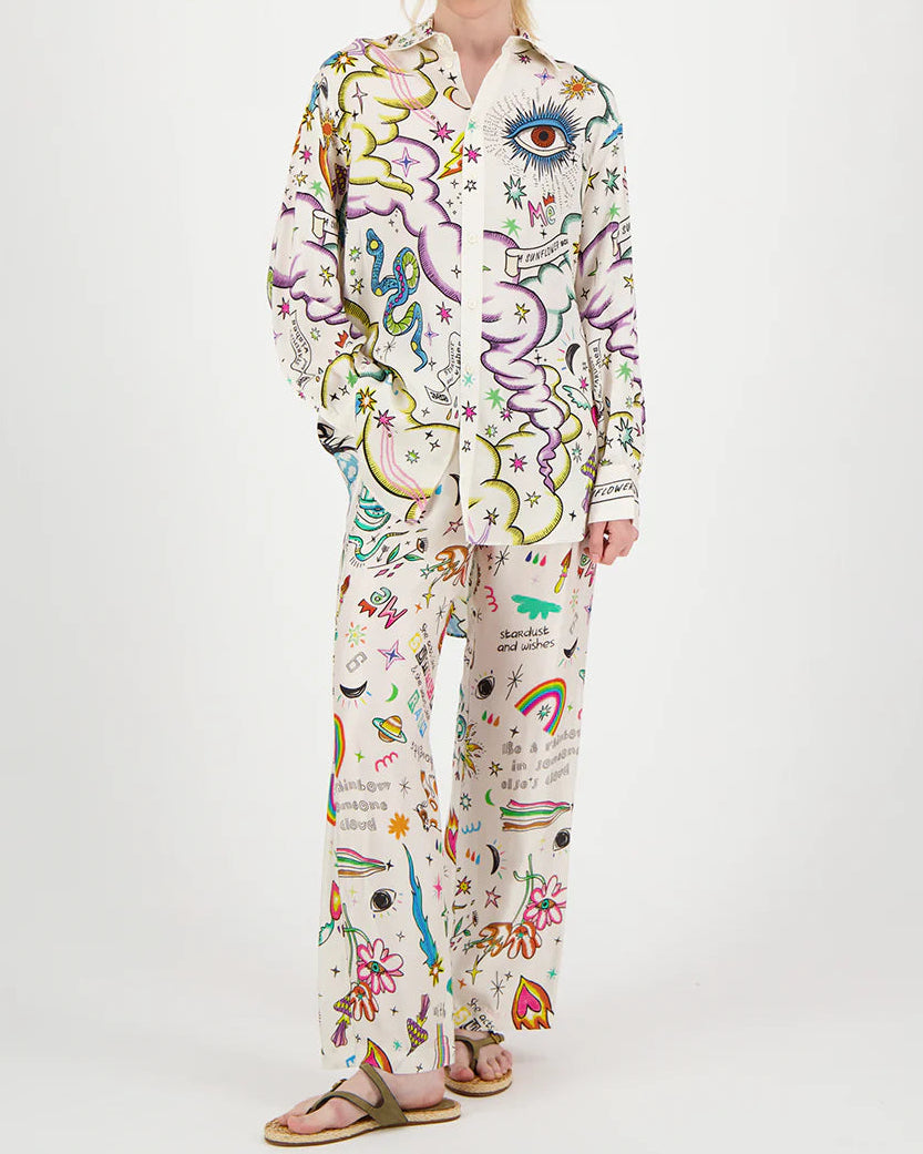 A person models ME369 Bailey Pants—loose-fitting pajama pants with playful, colorful prints of eyes, rainbows, stars, clouds, hearts, and flowers—against a plain white background, showcasing true dopamine dressing by ME369.