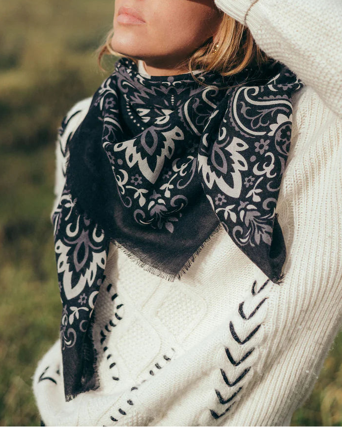 Wearing a cream knit sweater with black stitching and a Maison Lecomte Flament Bandana Scarf in black, featuring a white paisley pattern, a person stands outdoors in sunlight, gazing thoughtfully into the distance.