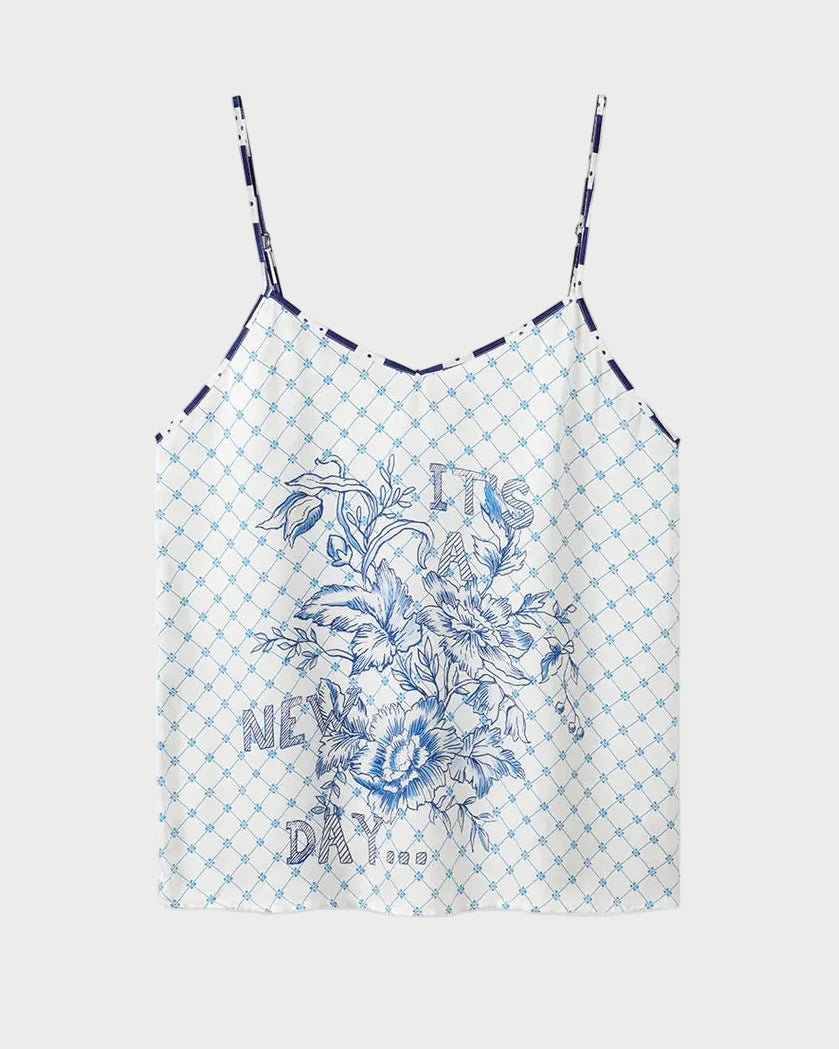 The ME369 Angelique Camisole is a white top with thin straps, showcasing a blue geometric check print and an intricate blue floral design on the front. Inspired by the original style, this camisole also features the phrase "IT'S A NEW DAY" printed in blue among the flowers.