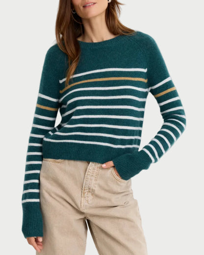 A woman wearing a dark teal knit sweater with horizontal white and mustard yellow stripes, paired with beige trousers, standing against a plain white background.