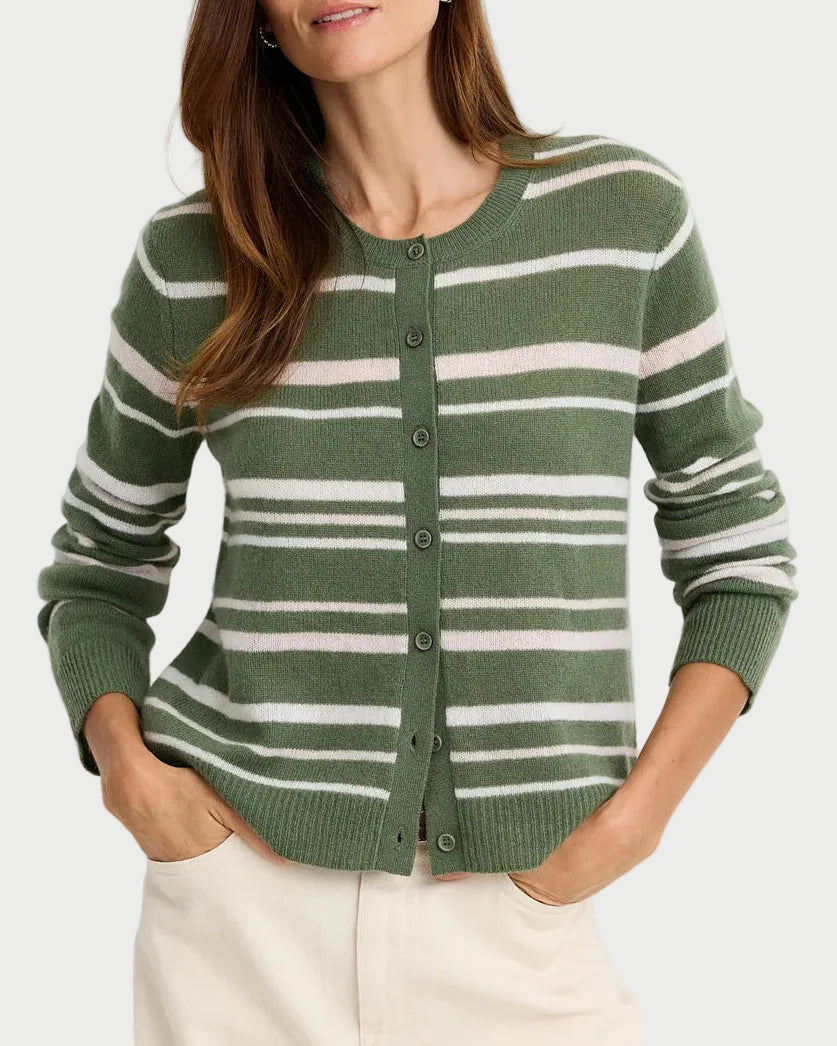 A woman wearing a green and white striped knit cardigan sweater with buttons, paired with cream-colored pants, standing against a plain light gray studio background.
