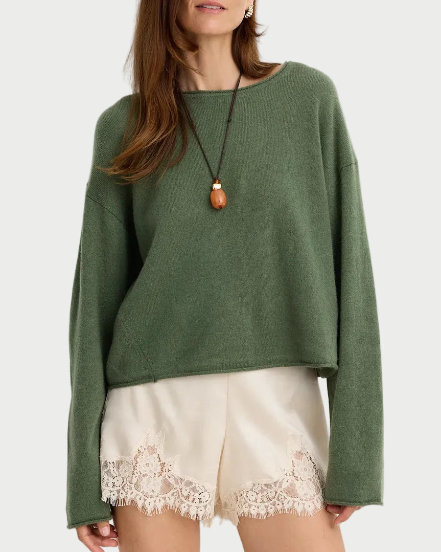 A woman modeling a sage green knit crewneck sweater paired with cream lace-trimmed shorts and a long pendant necklace, set against a plain light gray studio background.