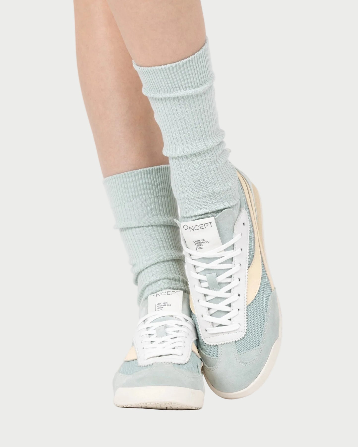 A person wearing light blue ribbed socks and pastel blue Oncept Reno Sneakers with cream accents stands with feet crossed on a light background, highlighting the Oncept Reno Sneaker’s effortless retro running vibe.
