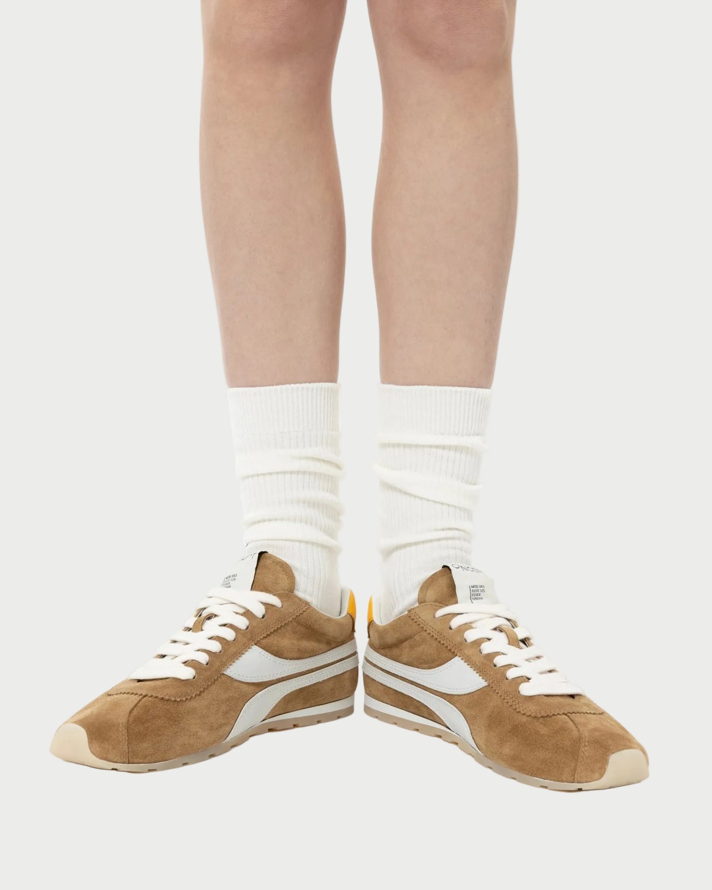 A person wears white socks and Oncept Essex Sneaker - Ginger by Oncept, featuring brown suede with white accents, sustainably crafted, standing on a light background. Only the lower legs and feet are shown.