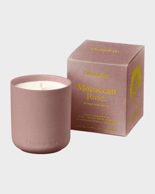 A pink candle in a matching holder sits beside its box, labeled "Osmology Moroccan Rose 9.8oz Candle," adorned with gold text and decorative elements. Infused with essential oils, the "Osmology" brand name is elegantly displayed against a plain white background.