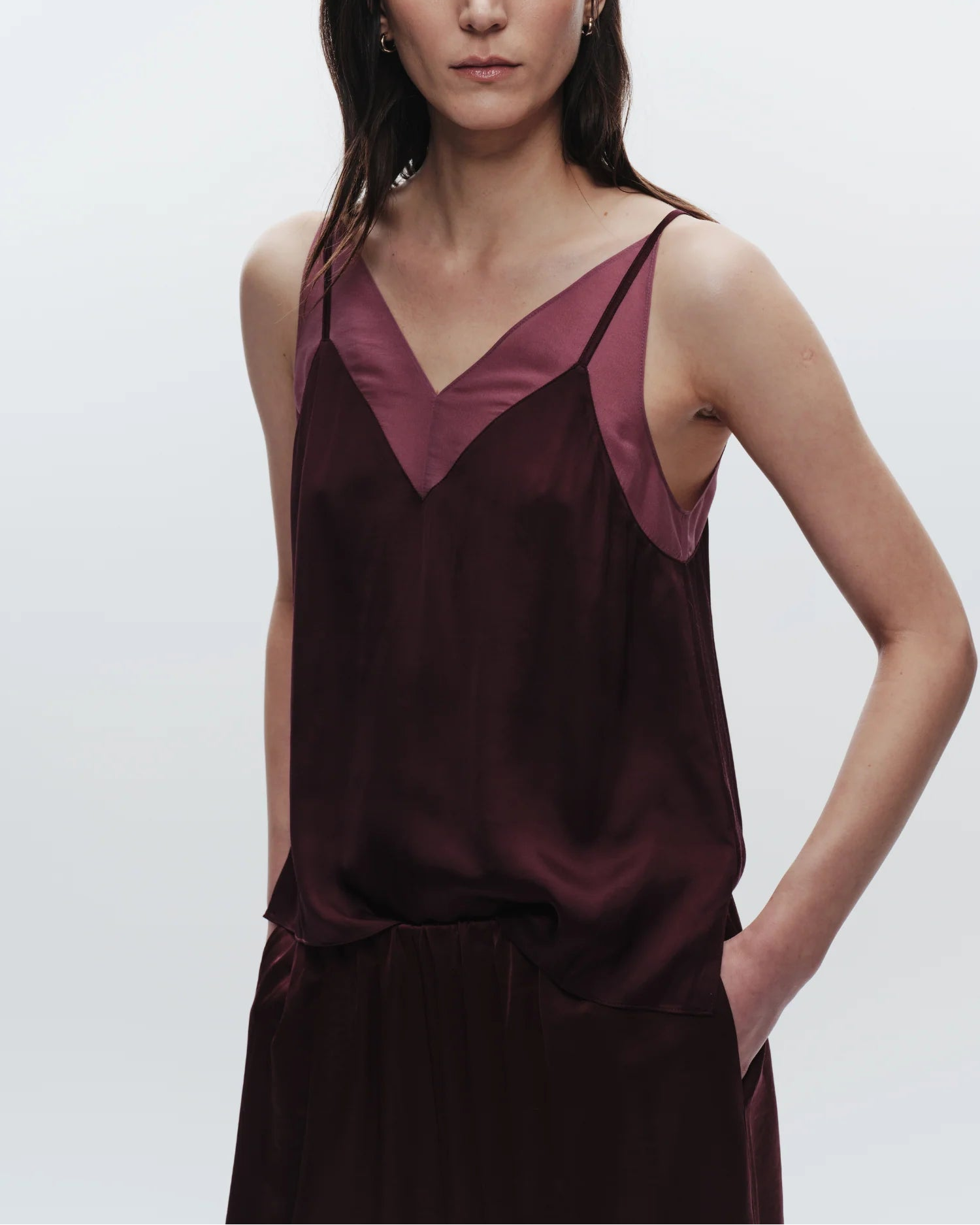 A woman wears the Saint Art Odin Camisole in Grape by Saint Art NY, a sleeveless satin top with a V-neckline and matching pants, standing against a plain light background with her left hand in her pocket.