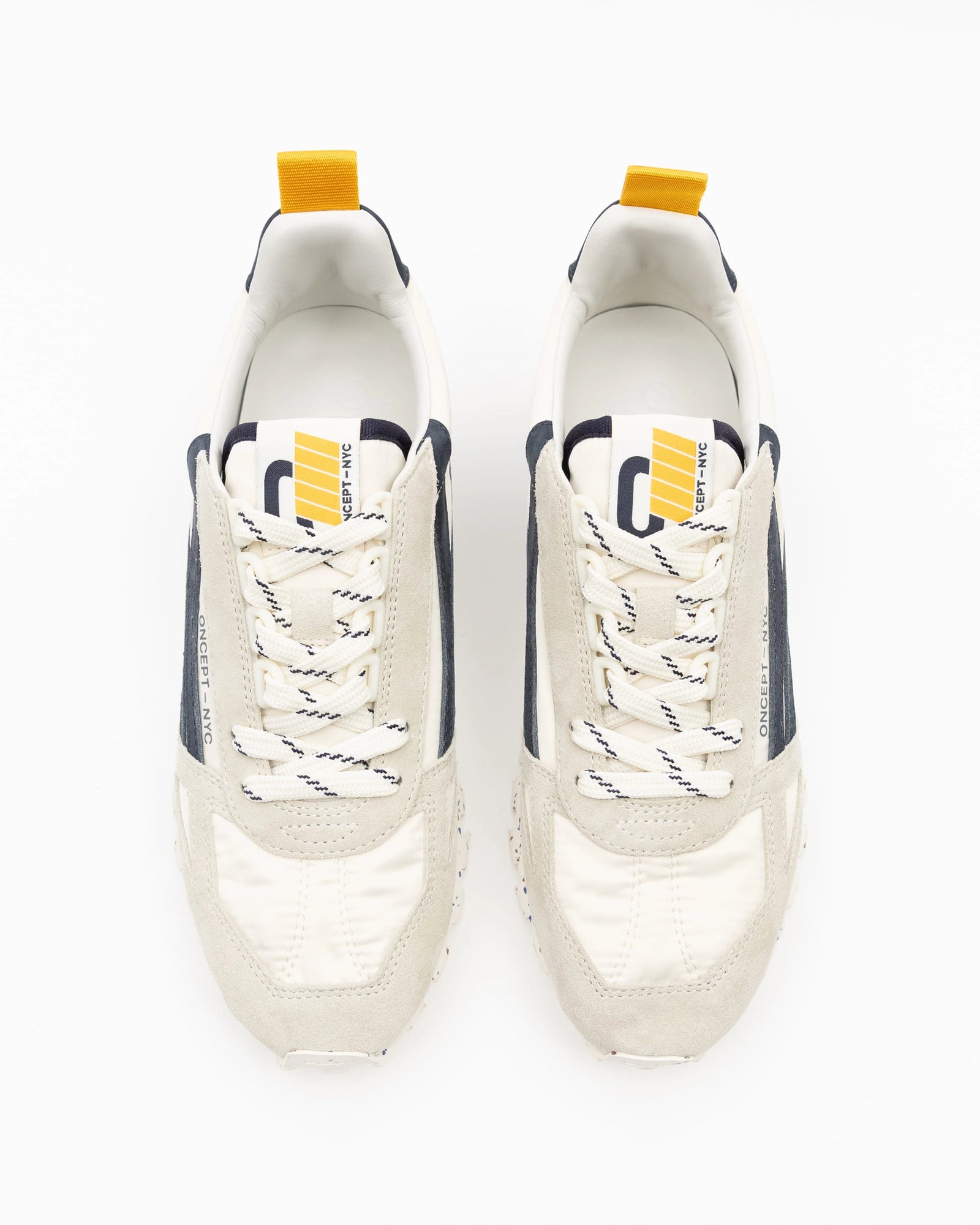 The Oncept Toronto Sneaker by Oncept features beige suede toes, white and navy panels, thick white laces with black accents, and yellow heel pull tabs—crafted from sustainable materials and perfect for travel, seen from above on a white background.