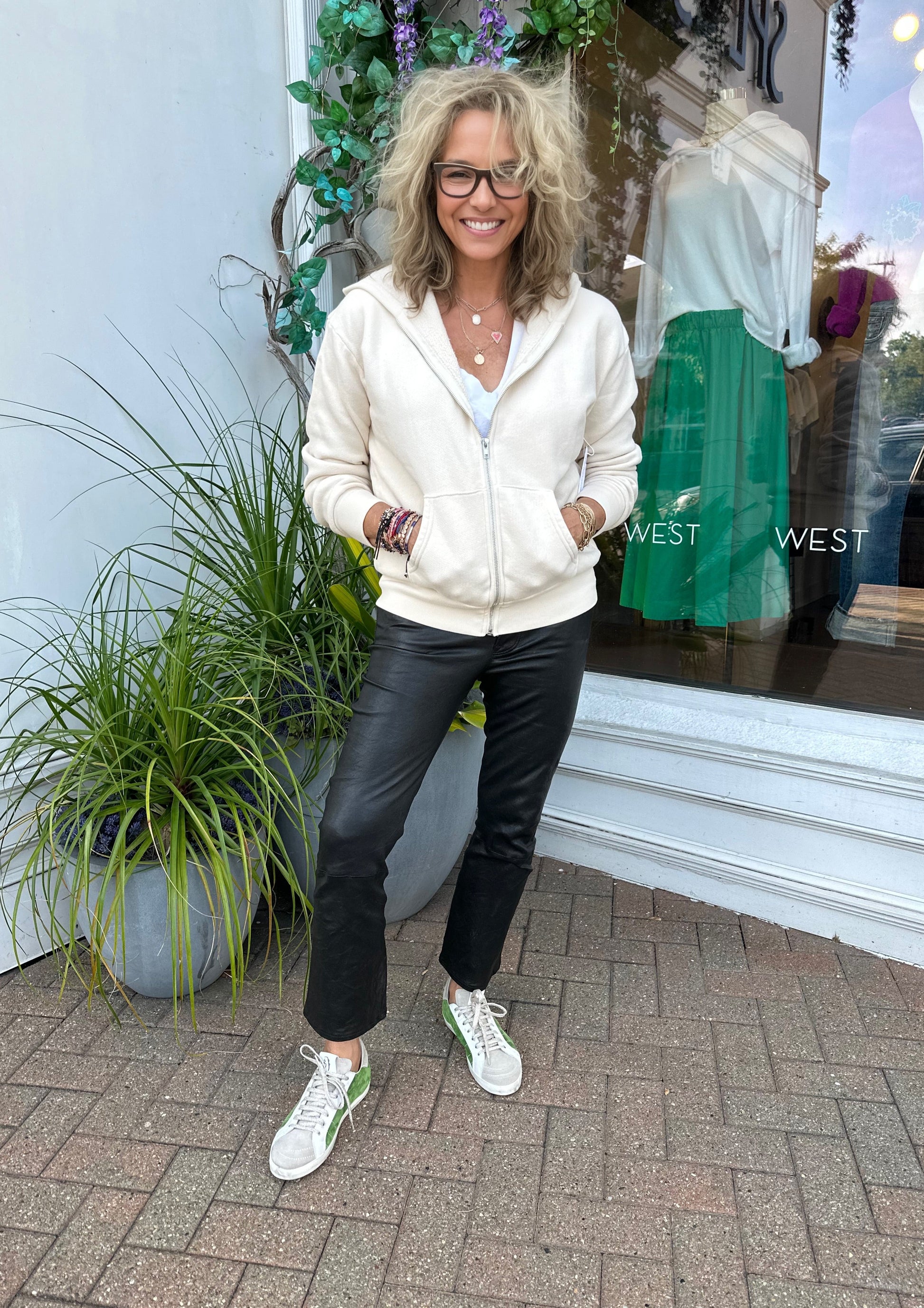 A woman with curly blonde hair, wearing glasses, a white hoodie, Le Crop Mini Boot Leather Pants—a true wardrobe essential by Frame—and white sneakers with green accents, smiles while standing outside a store with a large window. She is standing on a brick walkway next to a potted plant.