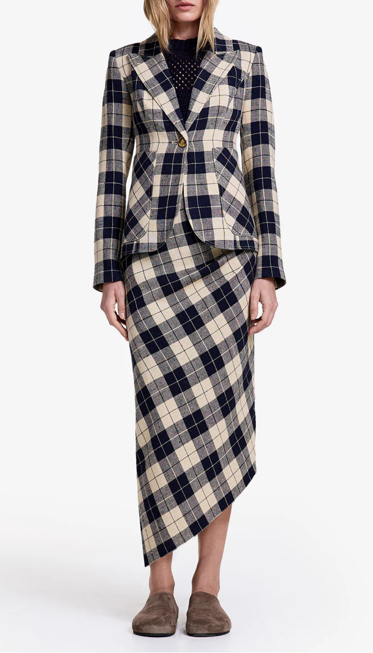 A person wears the Smythe Patch Pocket Duchess Plaid Blazer in navy, styled with a matching asymmetrical skirt, a dark top, and brown slippers, standing against a plain white background.