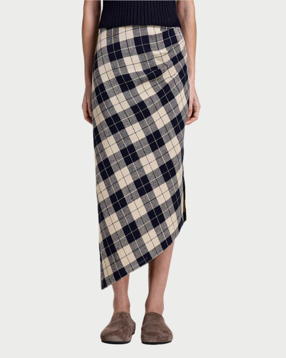 A person wears the Smythe Asymmetrical Plaid Skirt, a high-waisted midi with navy blue and beige plaid, asymmetric hem, and side slit, paired with brown slippers.