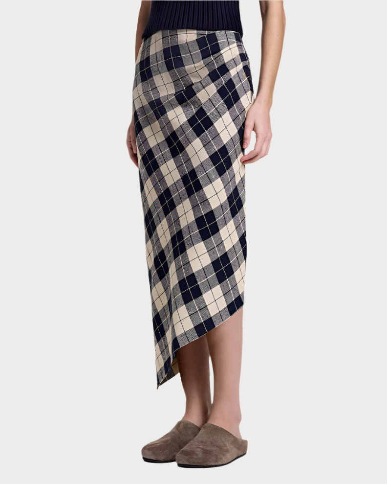 A person wears Smythe's Asymmetrical Plaid Skirt with a black sleeveless top and brown slippers, standing against a plain light gray background.