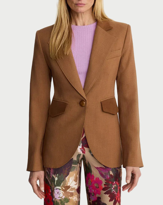 A blonde model wearing a camel brown blazer over a light purple top and floral print trousers, photographed against a plain light gray studio background.