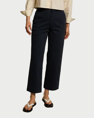 A person stands against a plain light background in the Perfectwhitetee Pia Twill Pant by perfectwhitetee—a high-waisted black utility trouser—paired with a beige wide-cuff long-sleeve top and black flip-flop sandals.