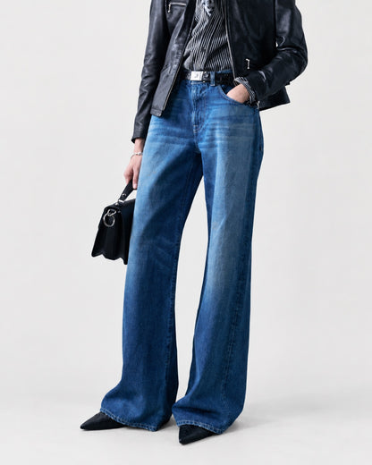 A person wearing R13 D’Arcy Flare Jean medium wash denim jeans, a black leather jacket, and black pointed-toe boots, holding a black handbag.