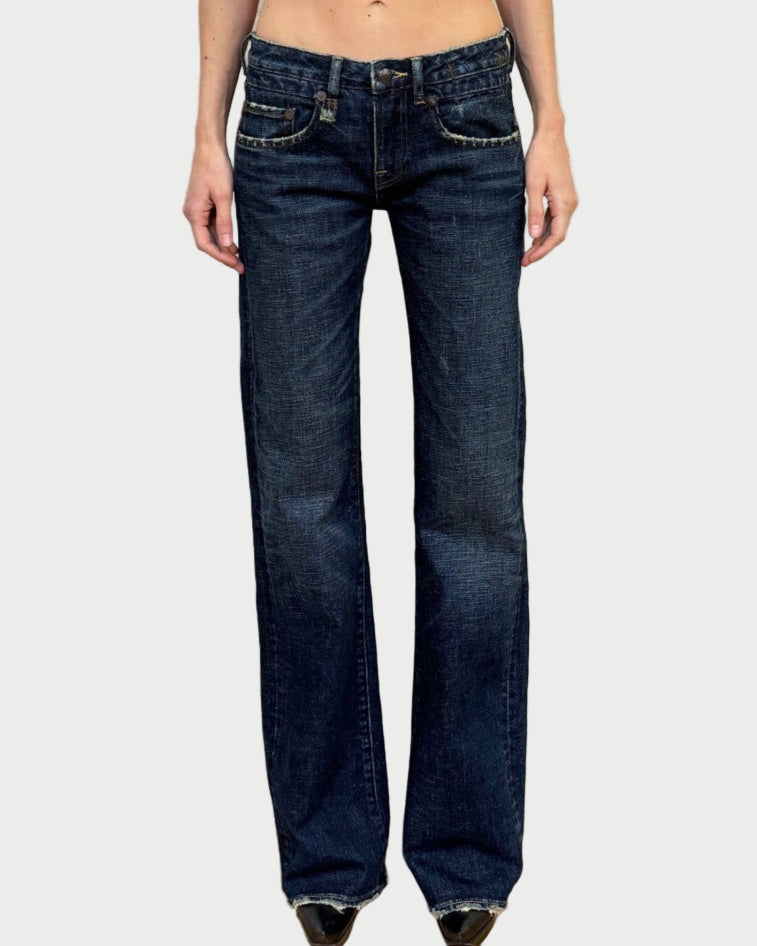 Model wearing R13 Boy Flare Jeans in dark blue with black pointed-toe shoes, standing against a white textured wall and wooden floor. Image cropped at the waist with face not shown. Brand: R13.