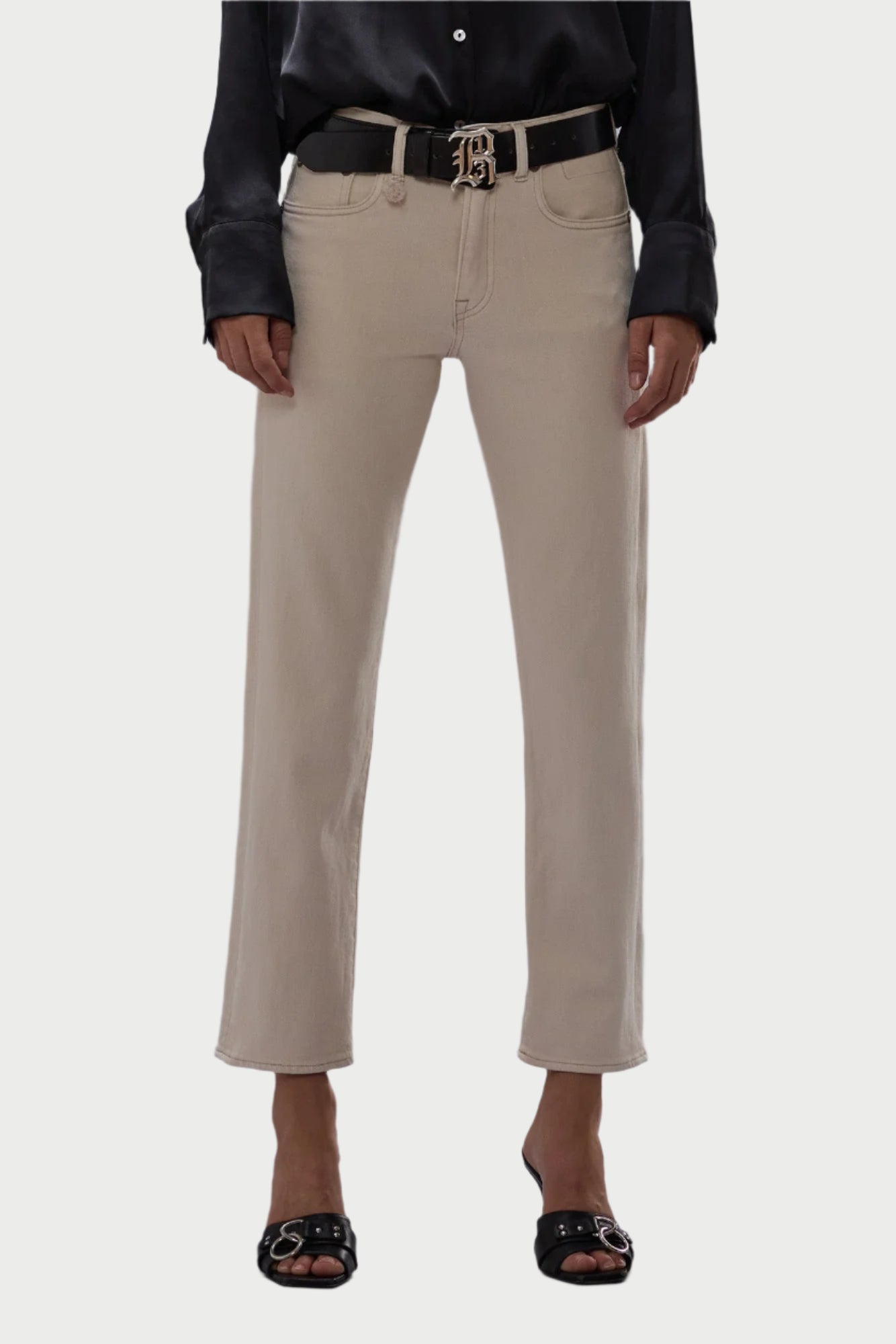 A person in a black long-sleeve shirt and beige high-waisted pants styled like R13's Romeo Jean in Ecru, complemented by a black belt with a decorative buckle and Japanese comfort stretch black open-toe sandals. The background is light gray.