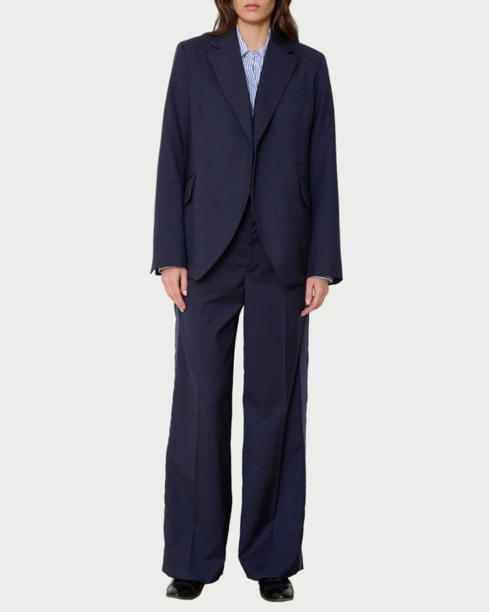 A person stands facing forward in the WE-AR4 Smoking Blazer by WE-AR4, a navy blue sustainable blazer made from upcycled virgin wool, paired with wide-leg trousers, black shoes, and a blue striped shirt against a white background.