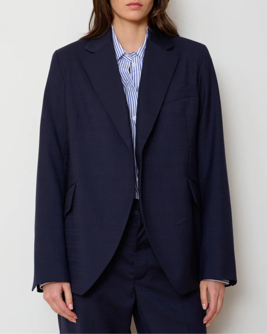 A person wearing the WE-AR4 Smoking Blazer by WE-AR4 with matching trousers and a blue and white striped button-up shirt stands against a plain light-colored background.