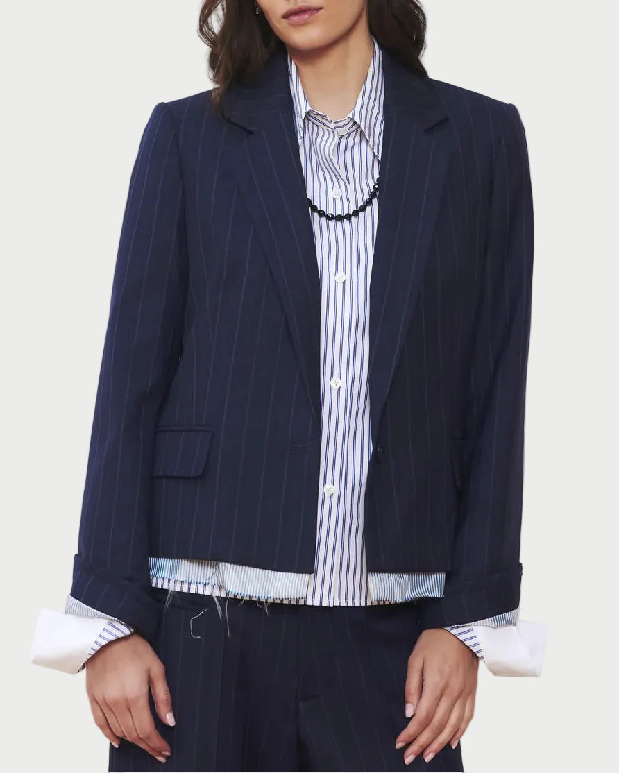 A woman wears the WE-AR4 The Hutton Blazer in navy pinstripe by WE-AR4 over a blue and white striped button-up shirt, with the shirt’s cuffs and hem peeking below the blazer. The look is finished with a black necklace.