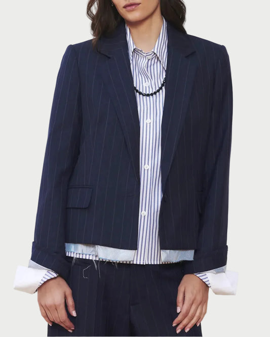 A woman wears the WE-AR4 The Hutton Blazer in navy pinstripe by WE-AR4 over a blue and white striped button-up shirt, with the shirt’s cuffs and hem peeking below the blazer. The look is finished with a black necklace.