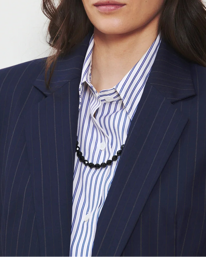 A person wearing the WE-AR4 The Hutton Blazer, a modern tailored navy pinstripe blazer by WE-AR4, over a blue and white striped shirt and a black beaded necklace.