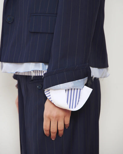 A person wearing the WE-AR4 The Hutton Blazer, a dark pinstripe suit jacket by WE-AR4, layered over frayed-edged shirts. Striped shirt cuffs extend past the blazer sleeves. Only the torso and one hand are visible, showcasing a distinctive, contemporary silhouette defined by the modern tailoring of The Hutton Blazer.