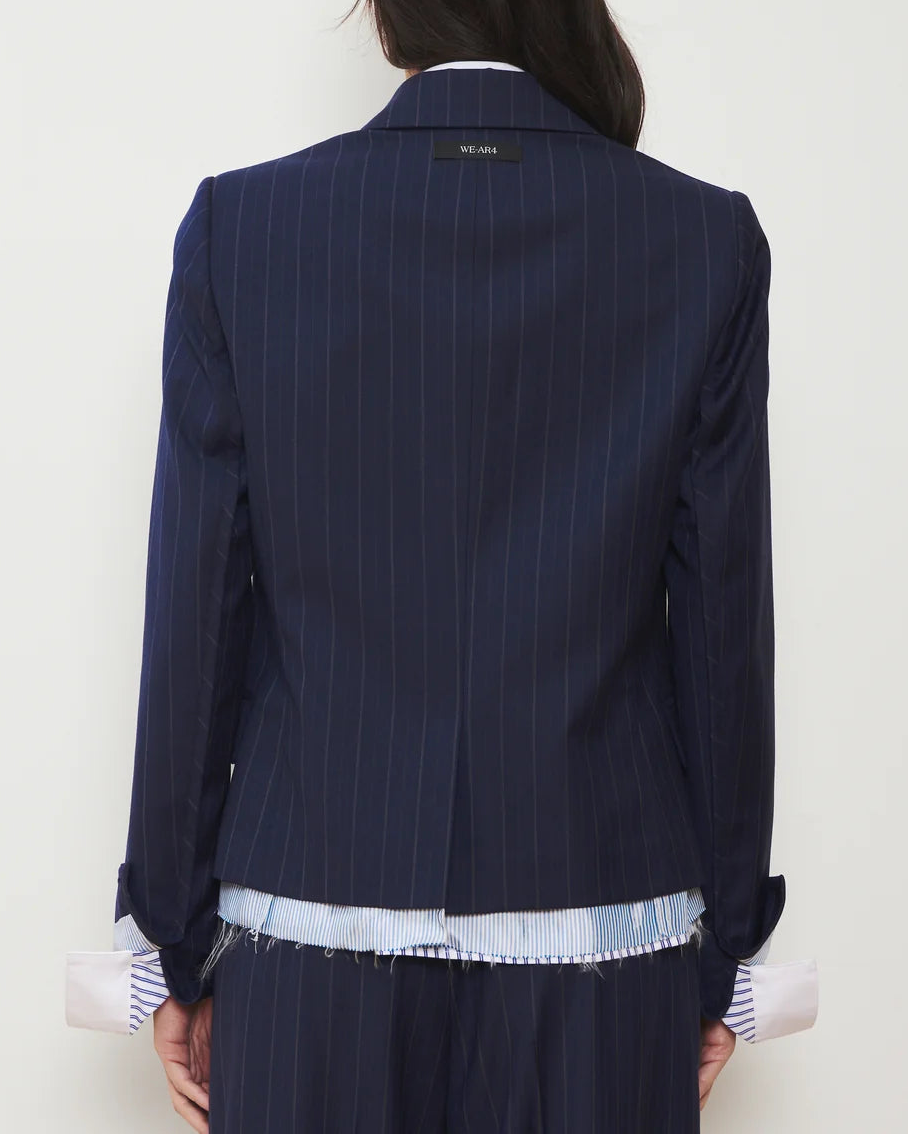A person seen from behind is wearing the WE-AR4 The Hutton Blazer by WE-AR4, a modern tailored jacket in navy blue pinstripes with a label at the neck. The blazer is layered over a blue and white striped shirt featuring frayed edges and pink cuffs peeking out.