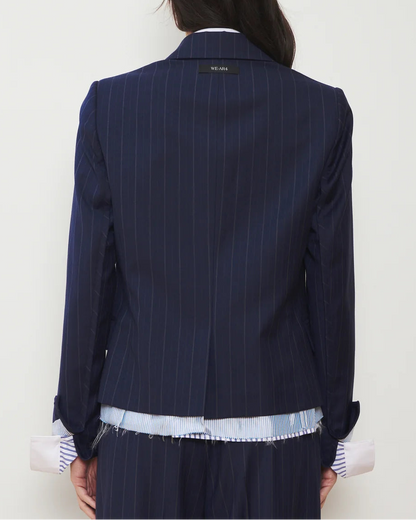 A person seen from behind is wearing the WE-AR4 The Hutton Blazer by WE-AR4, a modern tailored jacket in navy blue pinstripes with a label at the neck. The blazer is layered over a blue and white striped shirt featuring frayed edges and pink cuffs peeking out.