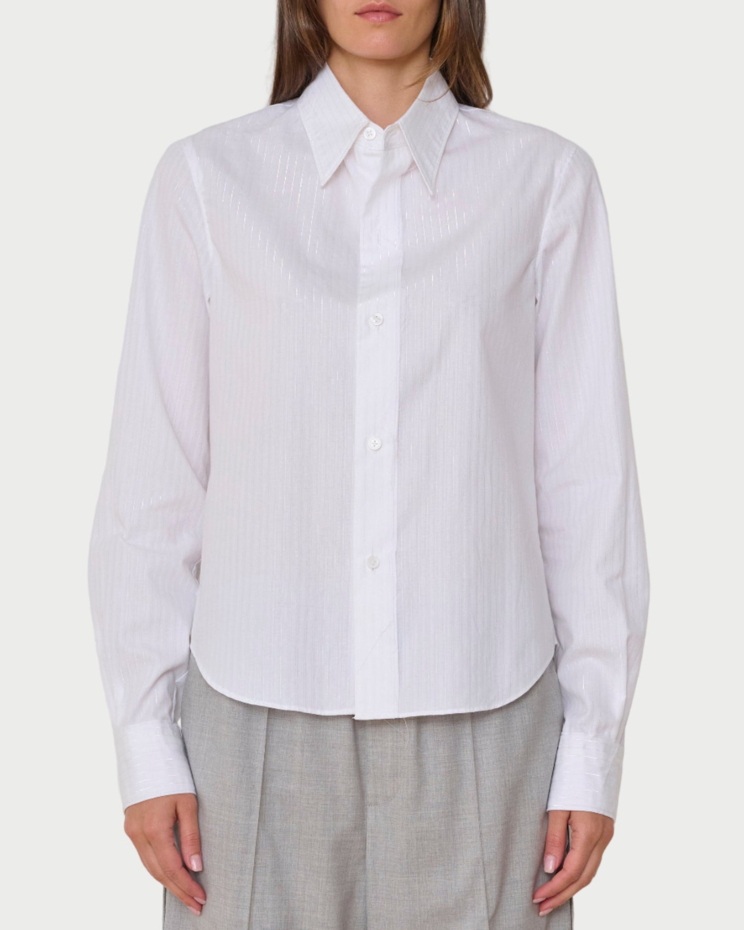 A person wears the WE-AR4 Rebel Shirt White by WE-AR4, a collared cotton poplin shirt with light vertical stripes, paired with light gray trousers, shown from the front against a plain background.