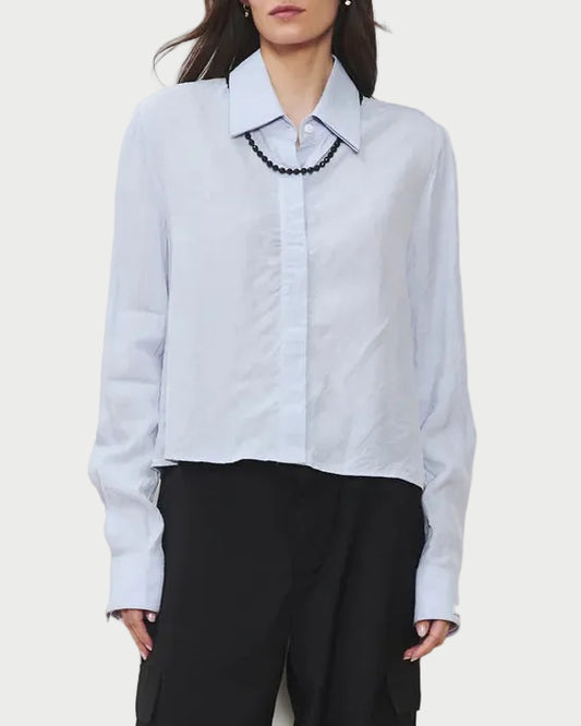 A woman wearing the WE-AR4 Campbell Shirt—a light blue modern button-down with long sleeves—styled with a black beaded necklace and black pants, standing against a plain white background.