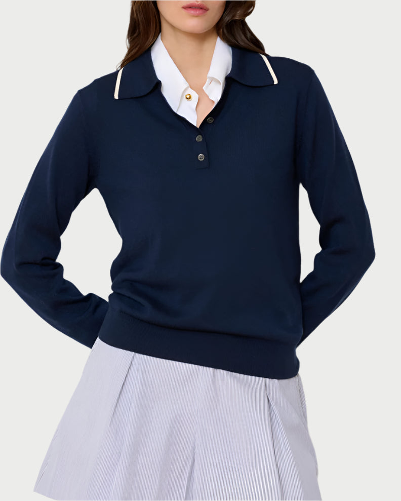 A woman wears the WE-AR4 Varsity Polo Sweater in navy blue over a white button-up shirt and light blue pleated skirt, standing with her hands behind her back against a plain light background.