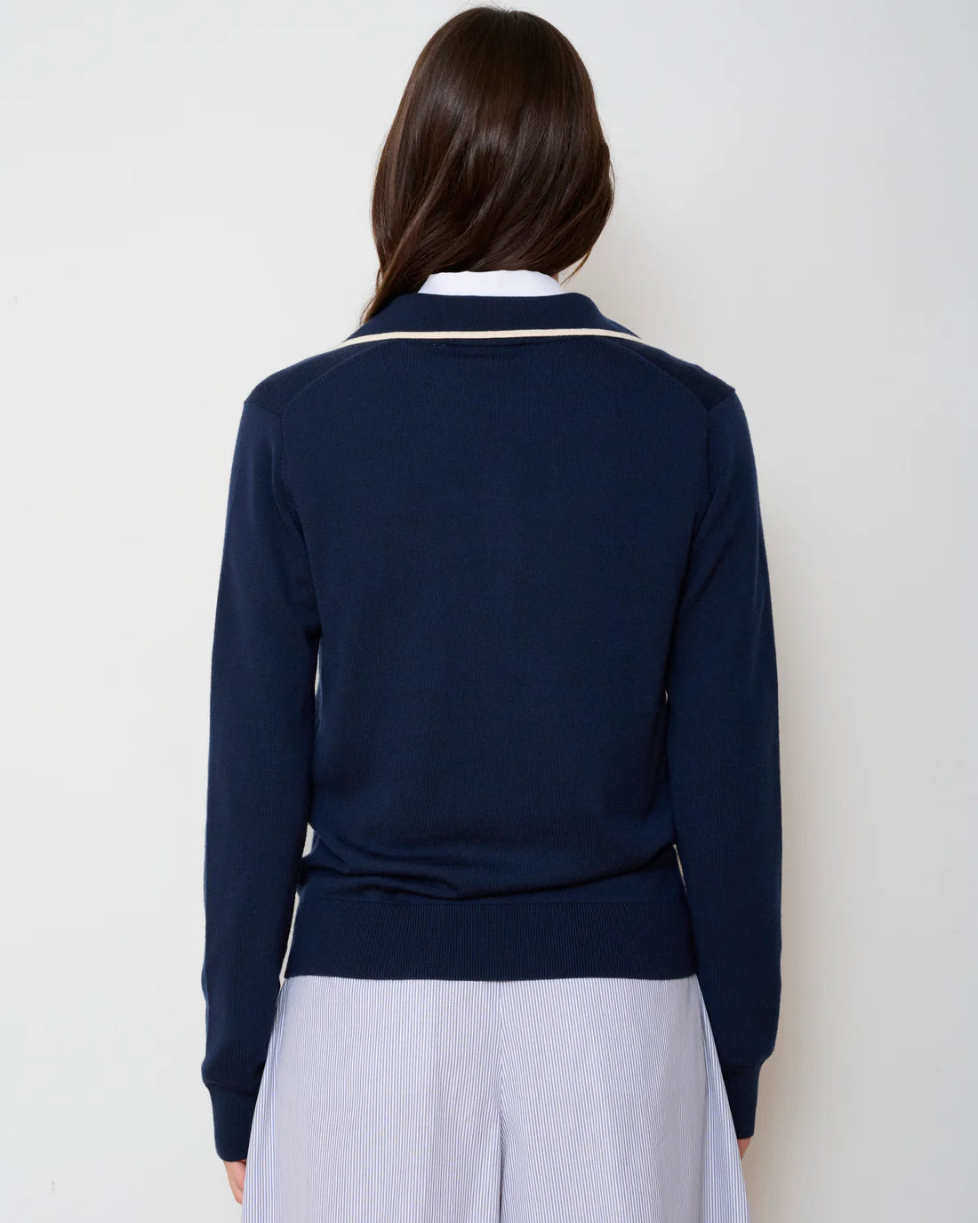 A person with long brown hair faces away from the camera, wearing the WE-AR4 Varsity Polo Sweater in navy over a white collared shirt and light blue skirt. The background is plain and light-colored.
