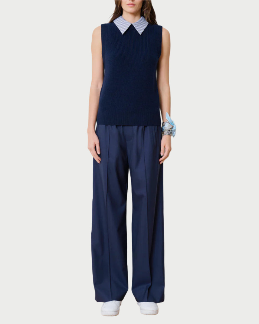 A person wears a sleeveless navy sweater crafted from upcycled Italian wool over a white collared shirt. The look is completed with WE-AR4 Prep Trousers and white sneakers, accented by a blue scarf wrapped around the wrist against a plain white background.