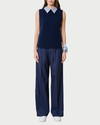 A person wears a sleeveless navy sweater crafted from upcycled Italian wool over a white collared shirt. The look is completed with WE-AR4 Prep Trousers and white sneakers, accented by a blue scarf wrapped around the wrist against a plain white background.