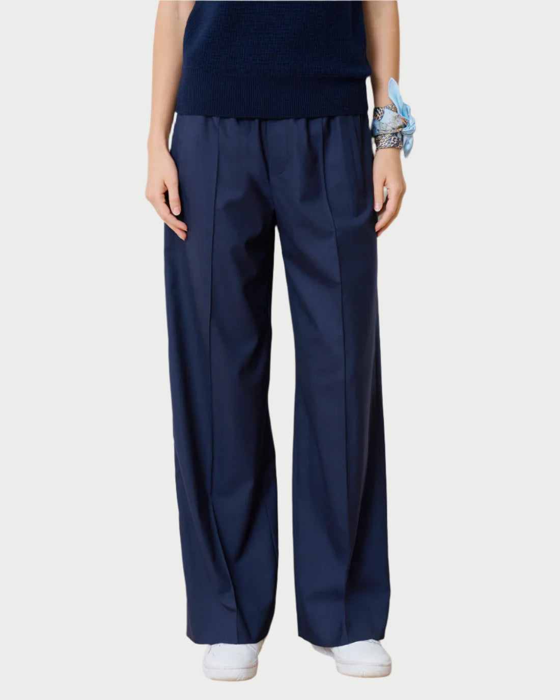 A person wears WE-AR4's navy blue wide-leg Prep Trouser made from upcycled Italian wool blend with a dark partial-sleeved top, has a blue scarf on their wrist, and white sneakers on a light background for an effortless look.