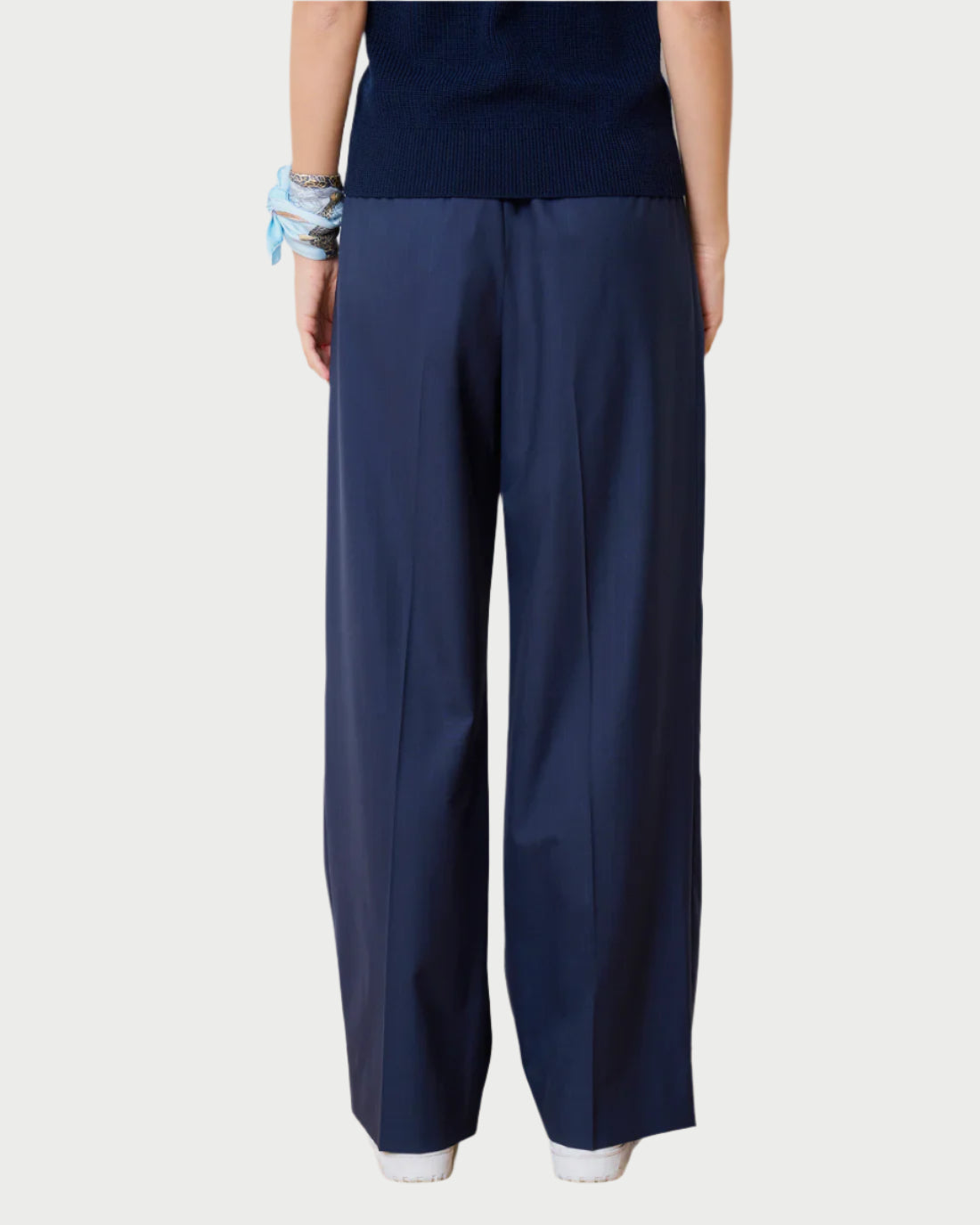 A person in WE-AR4’s Prep Trouser, wide-leg pants made from upcycled Italian wool blend, with a black top. They wear blue and silver bracelets on their left wrist. The plain light gray background captures the brand's chic style.