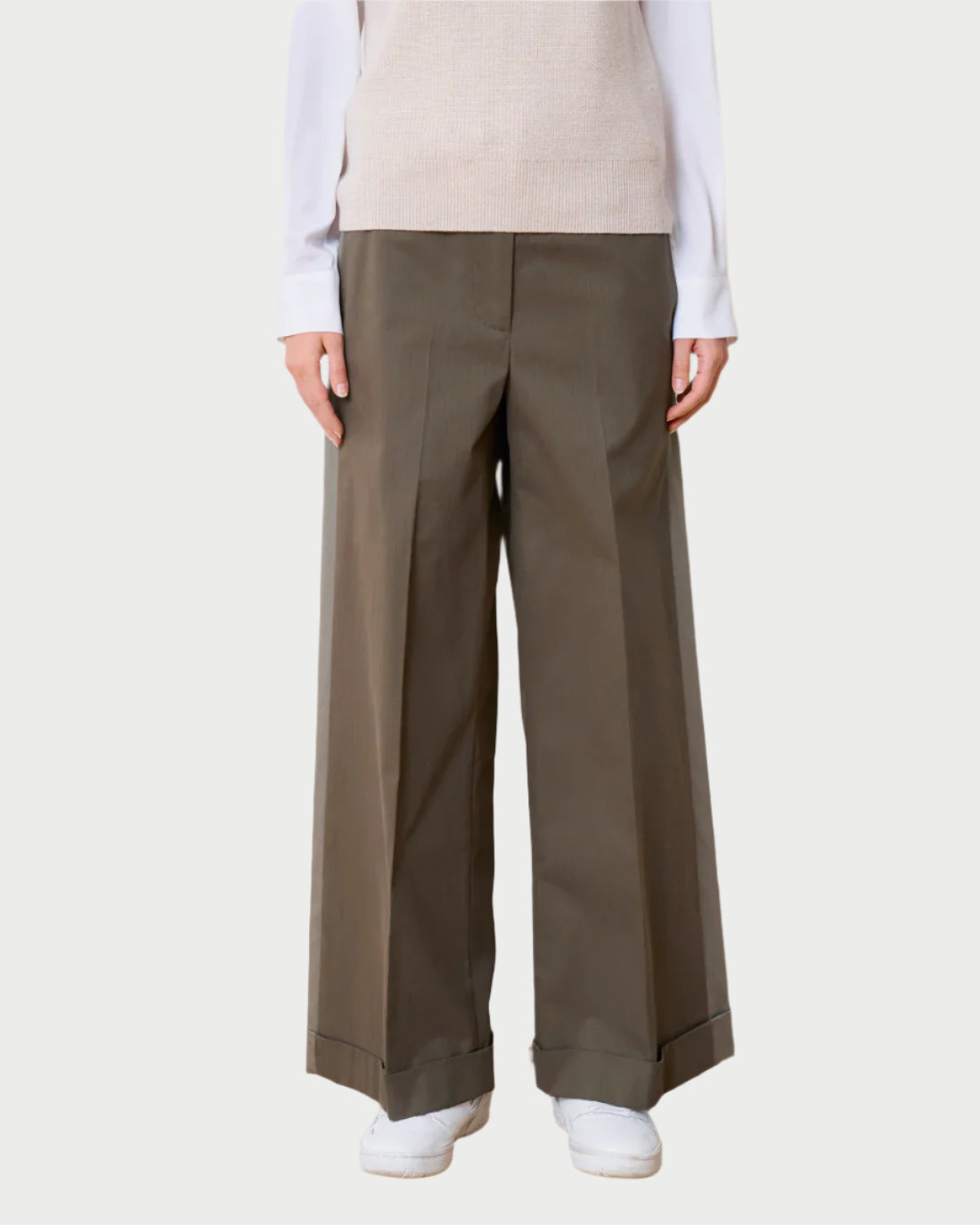 Against a light backdrop, someone dressed in a white long-sleeve shirt and beige sleeveless sweater sports WE-AR4's wide-leg olive green Field Trouser, crafted from upcycled cotton with prominent pleats, paired with white sneakers.