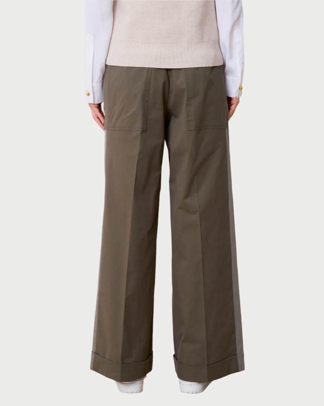 A person is shown from behind wearing WE-AR4's khaki Field Trouser, crafted from upcycled cotton with large pockets and a cuffed hem. The outfit includes a light beige sweater and a white shirt with buttoned cuffs, all set against a neutral backdrop.