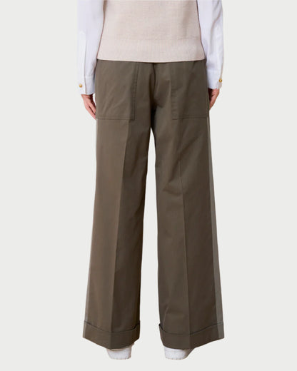 A person is shown from behind wearing WE-AR4's khaki Field Trouser, crafted from upcycled cotton with large pockets and a cuffed hem. The outfit includes a light beige sweater and a white shirt with buttoned cuffs, all set against a neutral backdrop.