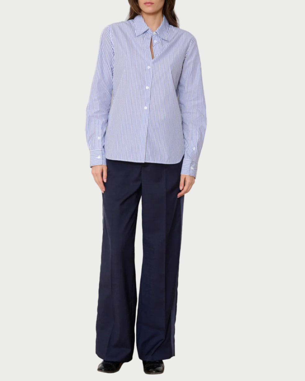 Woman wearing a blue and white striped shirt and navy pants against a white wall.