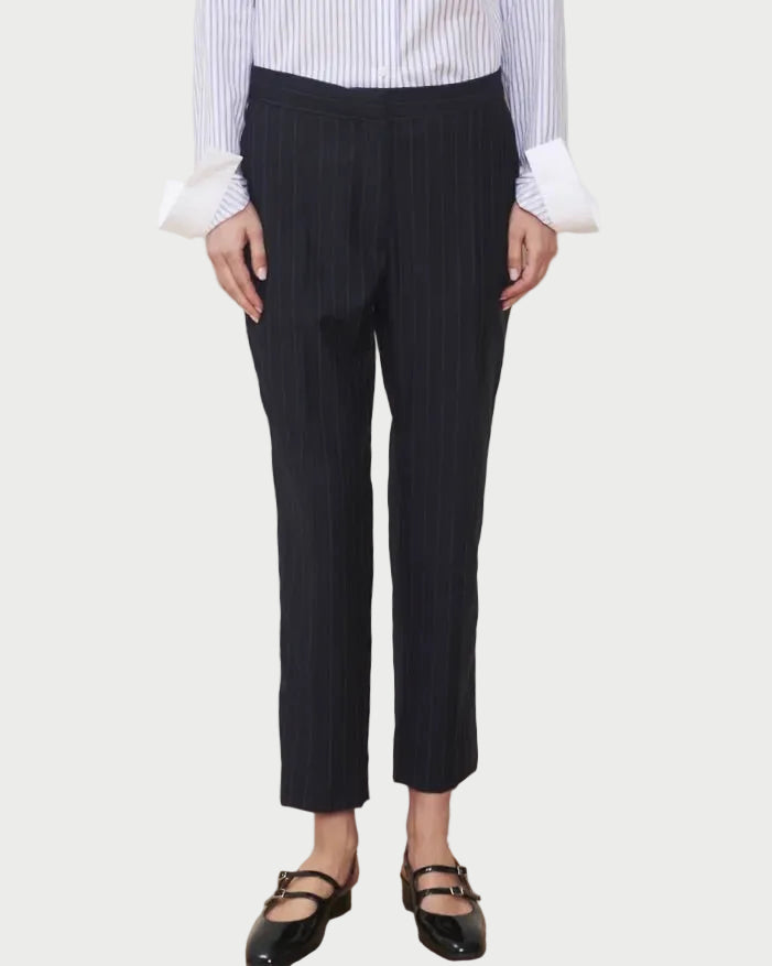 Wearing the WE-AR4 Windsor Trouser—a limited edition, cropped black pinstripe trouser with a tapered straight-leg silhouette—styled with a blue and white striped shirt featuring wide cuffs and black Mary Jane shoes, the person stands against a plain light background, their face not visible.