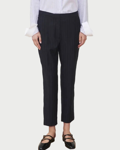 Wearing the WE-AR4 Windsor Trouser—a limited edition, cropped black pinstripe trouser with a tapered straight-leg silhouette—styled with a blue and white striped shirt featuring wide cuffs and black Mary Jane shoes, the person stands against a plain light background, their face not visible.