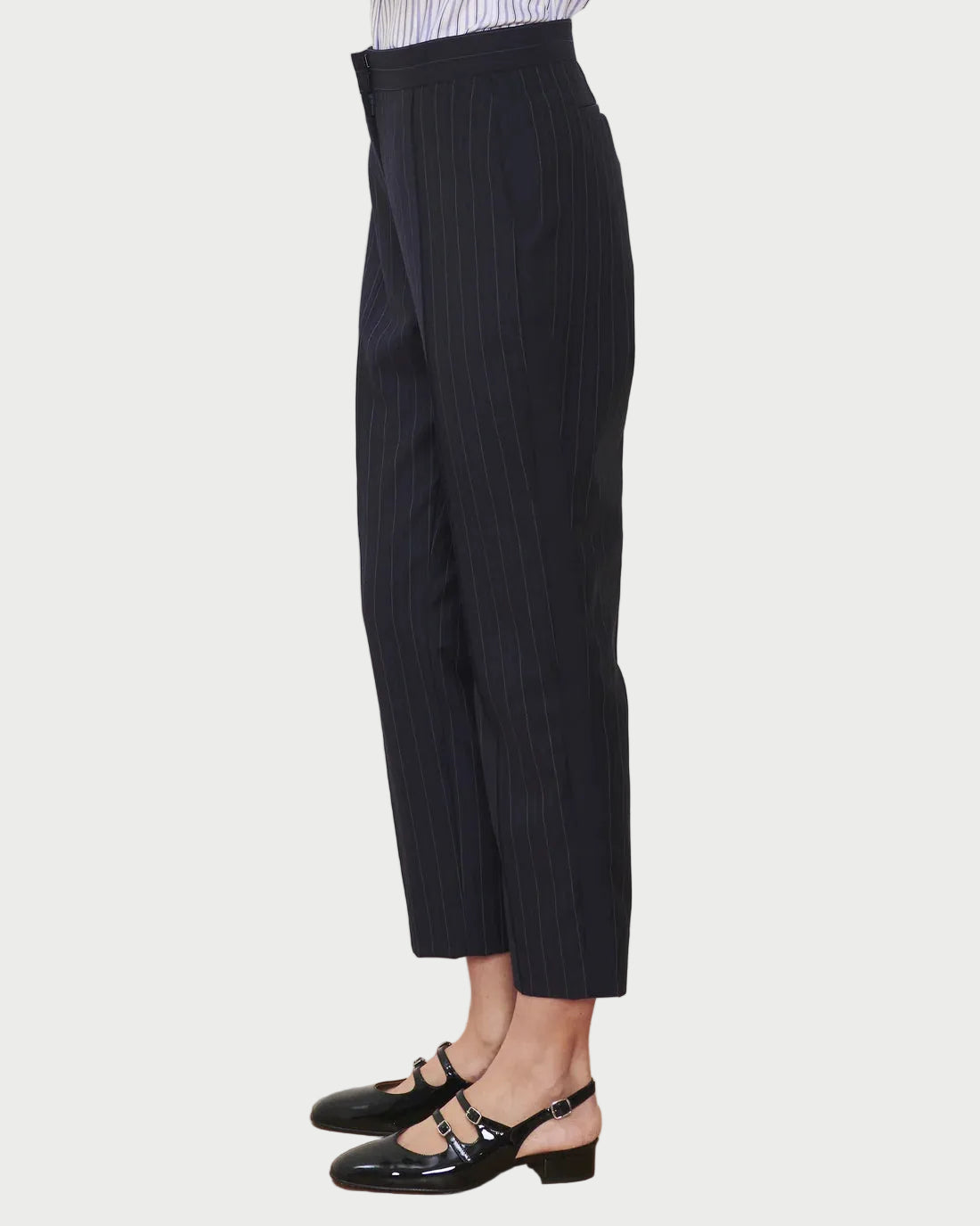A person wearing the WE-AR4 Windsor Trouser by WE-AR4, crafted from upcycled wool in a tapered straight-leg cut, paired with black patent Mary Jane shoes and standing sideways against a plain white background.