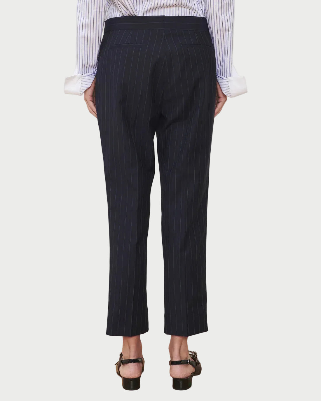 Wearing the WE-AR4 Windsor Trouser—a limited edition piece crafted from upcycled wool in a dark pinstripe—a person stands facing away, paired with a light striped shirt with rolled sleeves and black heeled sandals, all set against a white background.
